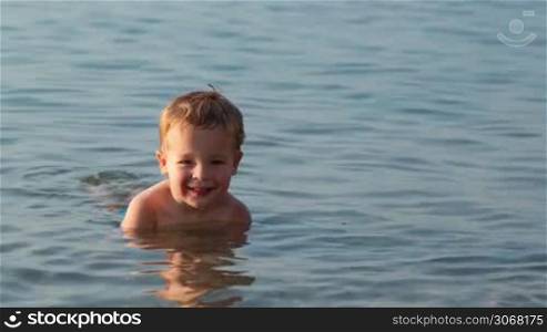 Smiling little boy ducking down in the shallow water at the seaside in a tranquil ocean on a summer day