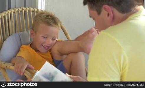 Smiling little boy is sitting in wicker chair and listening to the tale his father reading aloud.