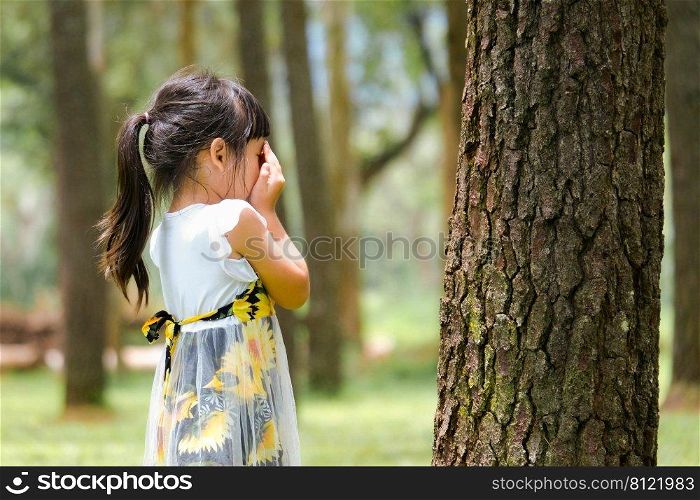 Smiling little girl covering her eyes with both hands, playing hide and seek standing beside a large tree. Cute little girl having fun outdoors in the park.