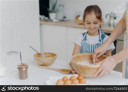 Smiling little girl helper holds big bowl, looks how mother is mixing eggs with flour, prepare tasty cake with chocolate, pose against cozy home kitchen interior, prepare food for all family