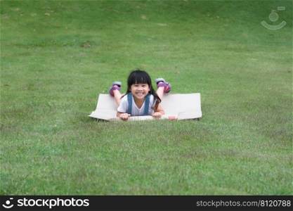 Smiling little girl lies prone on a cardboard box sliding down a hill at a botanical garden. The famous outdoor learning center of Mae Moh Mine Park, L&ang, Thailand. Happy childhood concept.