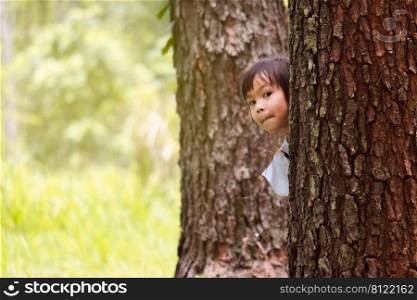 Smiling little girl standing behind a tree and looking at the camera. Adorable child having fun outdoors in the park. Childhood concept