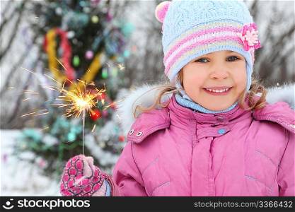smiling little girl wearing winter jacket is standing near christmass tree with bengal light.