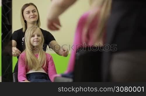 Smiling little girl with beautiful long blonde hair consulting with female master in hairdressing salon about hair cut. Cheerful child showing hairdresser necessary hair length to be cut off at barber shop.