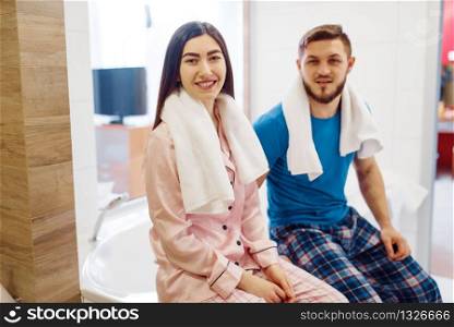 Smiling love couple in pajamas sitting in bathroom at home, morning hygienic procedure. Harmonious relationship in young family. Man and woman resting together in their house, carefree weekend. Couple sitting in bathroom, hygienic procedure