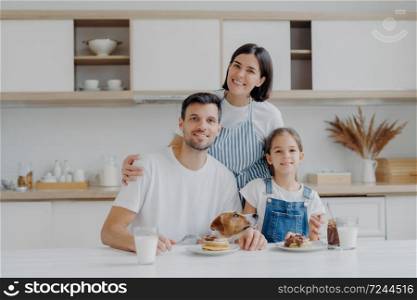 Smiling lovely mother and wife embrces with love daughter and husband, prepared delicious breakfast for them. Little child and father, their pedigree dog eat pancakes in kitchen. Friendly family