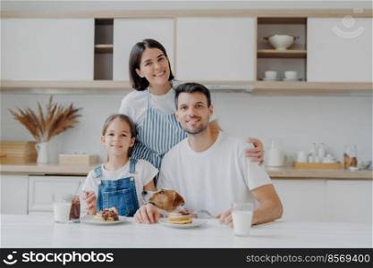 Smiling lovely mother and wife embrces with love daughter and husband, prepared delicious breakfast for them. Little child and father, their pedigree dog eat pancakes in kitchen. Friendly family