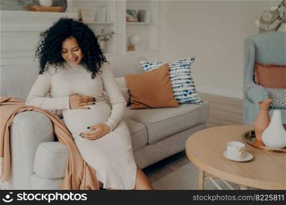 Smiling lovely pregnant mixed race woman in white dress with curly hair sitting on couch in front of round table and thinking about motherhood, happy to become mother. Pregnancy concept. Smiling beautiful pregnant mixed race woman sitting on couch and touching her belly
