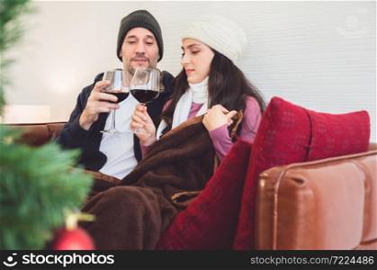 Smiling lovers sitting on sofa and clinking glasses of red wine in living room. Romantic winter holiday and Christmas eve together concept