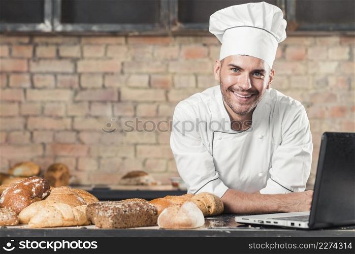 smiling male baker with different type baked breads laptop kitchen worktop