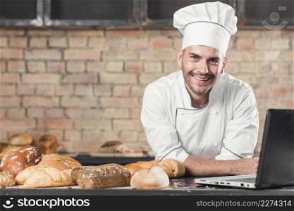 smiling male baker with different type baked breads laptop kitchen worktop