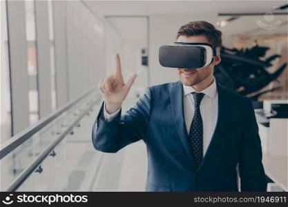 Smiling male business professional in suit wearing VR headset on head using virtual reality at workplace, businessman interacting with objects in cyberspace while standing in office. Future technology. Smiling business professional in suit wearing VR headset on head using virtual reality at workplace