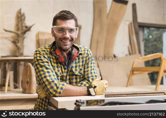 smiling male carpenter with ear defender around her neck standing his workshop