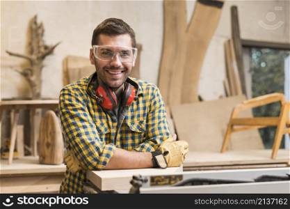 smiling male carpenter with ear defender around her neck standing his workshop