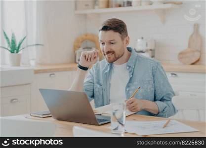 Smiling male employee using laptop at home sitting at kitchen table, looking at screen and making video call or watching webinar, writing notes in notebook. Distance studying and freelance concept. Smiling male employee using laptop at home while sitting at kitchen table