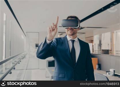 Smiling male entrepreneur using vr goggles for 3d visualization, pointing with finger, businessman wearing virtual reality glasses, interacting with digital world while standing in modern office. Smiling male entrepreneur using vr goggles for 3d visualization, pointing with finger