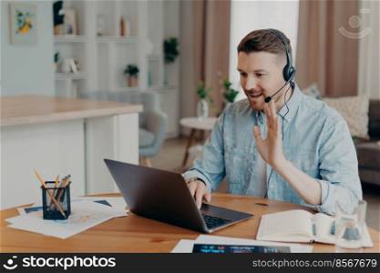 Smiling male freelancer working at home and using headset while looking at laptop screen and saying hello, waving to somebody and smiling, sitting in front of computer and having video call. Happy Caucasian guy waving hand during video call to colleague