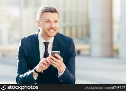 Smiling male manager with appealing look, dressed formally, chats with costumer, uses modern smart phone, sits against urban background, connected to wireless high speed internet. Technology