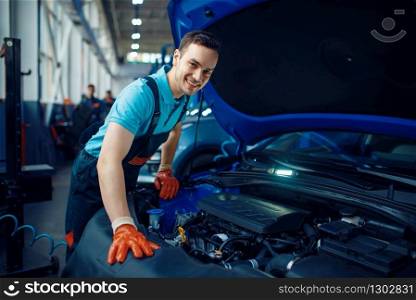 Smiling male worker in uniform checks vehicle engine, car service station. Automobile checking and inspection, professional diagnostics and repair. Smiling worker checks vehicle, car service station