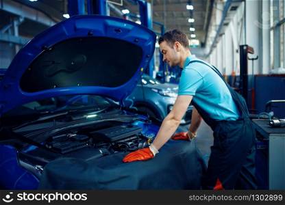 Smiling male worker in uniform checks vehicle engine, car service station. Automobile checking and inspection, professional diagnostics and repair. Smiling worker checks vehicle, car service station
