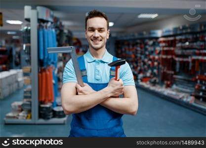 Smiling male worker in uniform holds hammer and corner in tool store. Choice of professional equipment in hardware shop, instrument supermarket. Worker holds hammer and corner in tool store