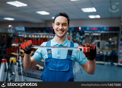 Smiling male worker in uniform holds measuring tape in tool store. Choice of professional equipment in hardware shop, instrument supermarket. Smiling worker holds measuring tape in tool store