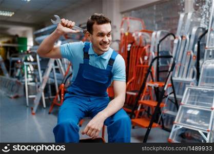 Smiling male worker in uniform holds wrench in tool store. Choice of professional equipment in hardware shop, instrument supermarket. Smiling male worker holds wrench in tool store