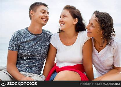 Smiling man and two young beautiful women sitting on beach, Looking against each other