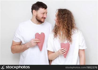 smiling man and woman in white t-shirts with red hearts in their hands lovingly looking at each other. couple in love on a white background. smiling man and woman in white t-shirts with red hearts in their hands lovingly looking at each other. couple in love on a white background.