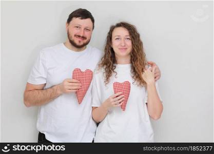 smiling man and woman in white t-shirts with red hearts in their hands lovingly looking at each other. couple in love on a white background. smiling man and woman in white t-shirts with red hearts in their hands lovingly looking at each other. couple in love on a white background.