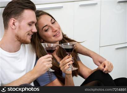 Smiling man and woman sitting on kitchen floor and holding glasses of cocktails. Romantic couple spending time together, having fun and enjoy drinking at home.