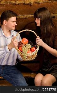smiling man and young woman with basket of fruit sitting on bench in wooden log hut, close up