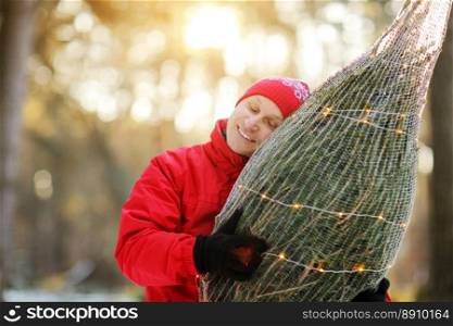 smiling man carrying freshly cut down christmas tree in forest. Young lumberjack bears fir tree on his shoulder in the woods. Irresponsible behavior towards nature, save forest, keep green concept. smiling man carrying freshly cut down christmas tree in forest. Young lumberjack bears fir tree on his shoulder in the woods. Irresponsible behavior towards nature, save forest, keep green concept.