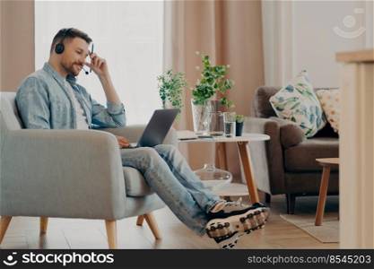 Smiling man freelancer looking at laptop screen during online video call with team while working remotely at his cozy home office. Lifestyle and freelance concept. Stylish happy man using laptop while working remotely at home