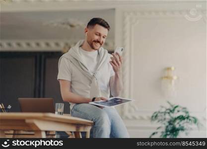 Smiling man freelancer wearing casual wear and earphones looking at phone and taking part in online meeting, casually sitting on desk and holding project documents in hand while working at home. Happy male freelancer looking at phone and smiling while sitting on work desk