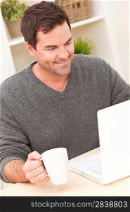 Smiling man in his thirties at home using his laptop computer drinkng tea or coffee