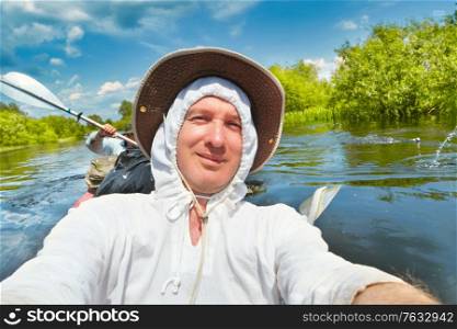 Smiling man in white hoodie and hat taking selfie photo on kayak during summer river adventure trip