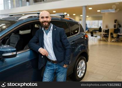 Smiling man poses at automobile in car dealership. Customer in new vehicle showroom, male person buying transport, auto dealer business. Smiling man poses at automobile in car dealership