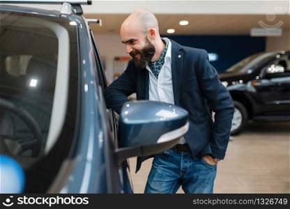 Smiling man poses at automobile in car dealership. Customer in new vehicle showroom, male person buying transport, auto dealer business. Smiling man poses at automobile in car dealership