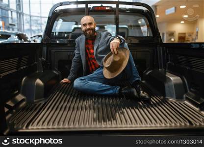 Smiling man poses in the back of new pickup truck in car dealership. Customer in vehicle showroom, male person buying transport, auto dealer business. Smiling man poses in the back of new pickup truck