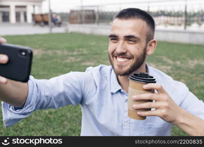 smiling man taking selfie while holding disposable coffee cup park