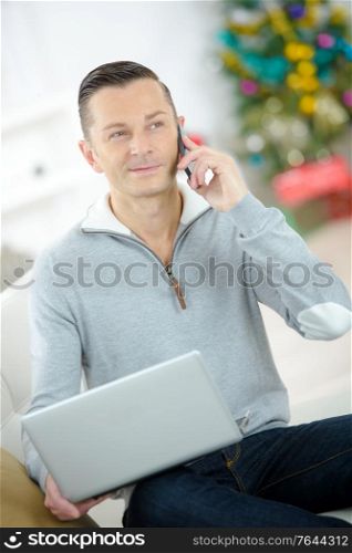 smiling man using laptop and phoning on sofa at home
