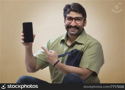 Smiling man wearing eyeglasses pointing towards a mobile phone which is held in other hand.