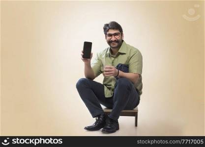 Smiling man wearing eyeglasses showing a mobile phone while holding a small pouch and a glass of tea.