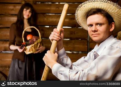 smiling man with pitchfork and in straw hat sitting on bench in wooden log hut, young woman with basket of fruit standing near wall on background, close up