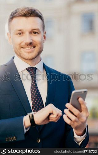 Smiling manager director with watch on arm, dressed elegantly, holds smart phone, reads positive news about his new project, poses outdoor, waits for colleague near office building. Job concept