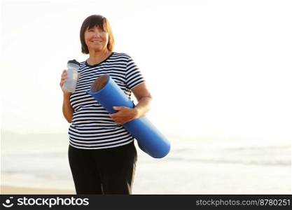 Smiling mature adult woman with bottle of water after exercising outdoors holding yoga mat outdoors on beach by sea on summer. Senior healthy lifestyle, vitality, healthcare concept. copy space.. Smiling mature adult woman with bottle of water after exercising outdoors holding yoga mat outdoors on beach by sea on summer. Senior healthy lifestyle, vitality, healthcare concept. copy space