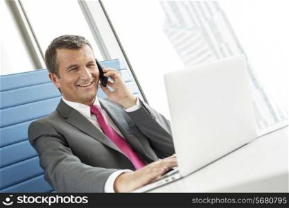 Smiling mature businessman talking on cell phone while using laptop in office