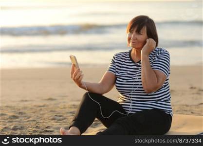 Smiling mature caucasian woman listening music in headphones looking at camera relaxing and resting on sandy beach by the sea at sunrise. Relaxation concept.. Smiling mature caucasian woman listening music in headphones looking at camera relaxing and resting on sandy beach by the sea at sunrise. Relaxation concept