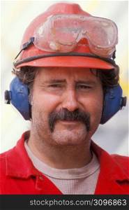 Smiling, mature construction worker with mustache in eyeglasses, hard-hat and headphones